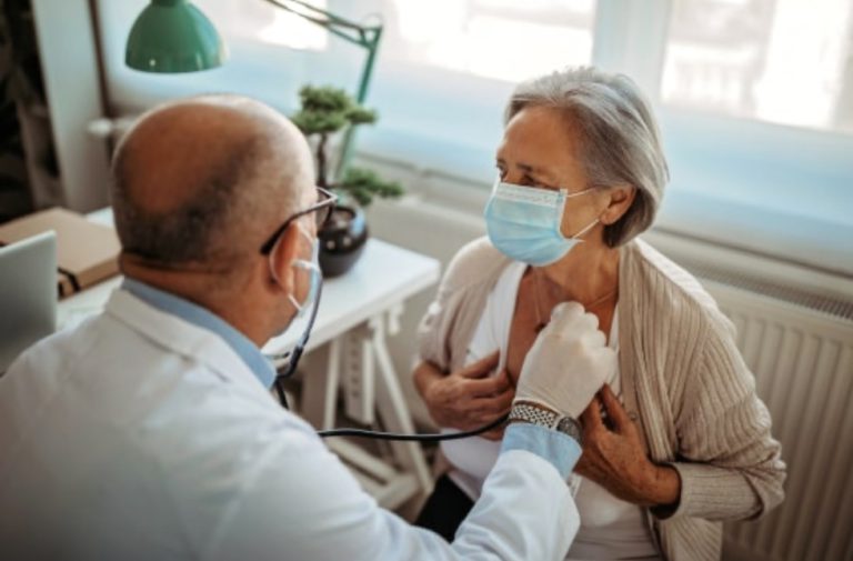 A male doctor is using a stethoscope to monitor an older woman's heart while wearing a face mask