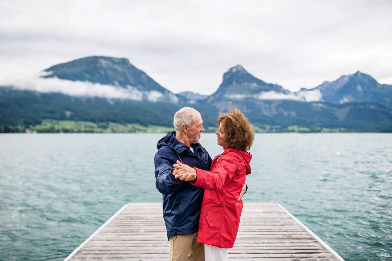 A senior couple are dancing on a jetty with a crystal blue lake and mountain peaks creating the perfect backdrop