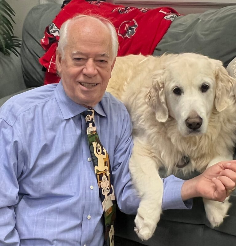 A senior gentleman wearing a blue shirt and a Wallace and Gromit tie sits next to dog.