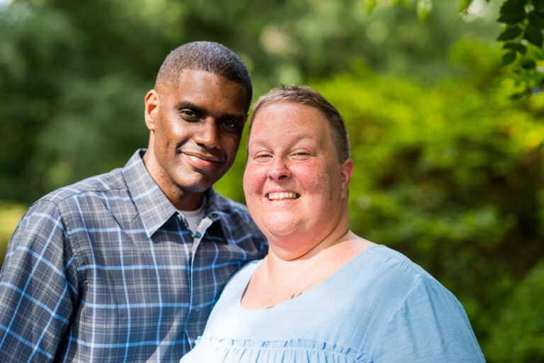 A couple posing for a photograph stood in front of background of green trees