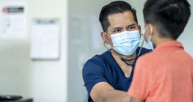 Male doctor wearing a mask and gloves during a checkup with a young, 8 year old boy because of the COVID-19 outbreak.