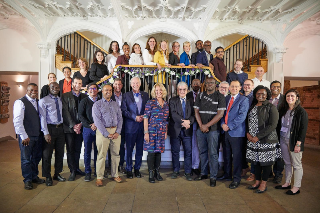 A large, diverse group of professionals poses on a staircase in a grand indoor setting with ornate white ceilings. Some stand on the stairs and others on the floor, all smiling towards the camera. The group are part of the emerging leaders programme.
