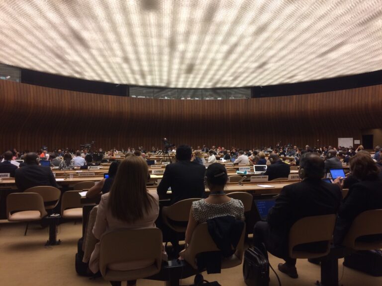 Wide-angle view of an international conference in session at the United Nations' Palais des Nations in Geneva, with delegates seated beneath a large, illuminated ceiling in a circular assembly hall.