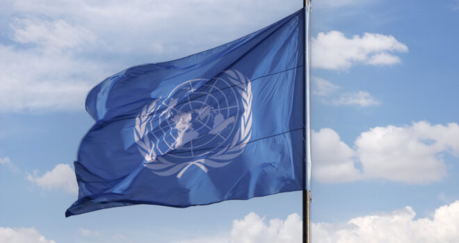 A blue United Nations flag blowing in the wind with a blue sky and clouds in the background