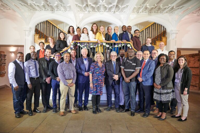 A large, diverse group of professionals poses on a staircase in a grand indoor setting with ornate white ceilings. Some stand on the stairs and others on the floor, all smiling towards the camera. The group are part of the emerging leaders programme.