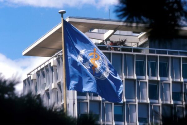 World Health Organization (WHO) flag waving in front of the WHO headquarters building in Geneva, Switzerland on a clear blue sky day.