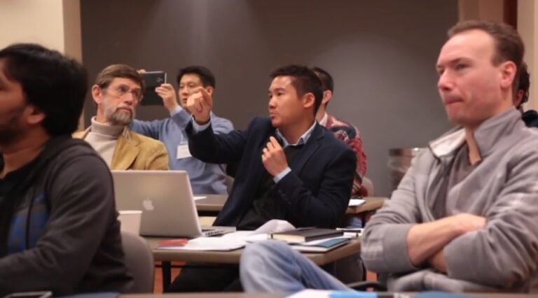 A man in a suit is speaking and gesturing with his hand while seated among a group of people in a classroom or seminar setting. Others around him are listening, taking notes, or recording with a phone. Laptops and notebooks are visible on the desks.