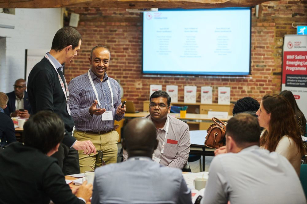 A candid photo of a professional networking or workshop session. A man is enthusiastically talking while standing, surrounded by seated and standing participants who are attentively listening. A presentation screen and banners indicating a leadership program are visible in the background.