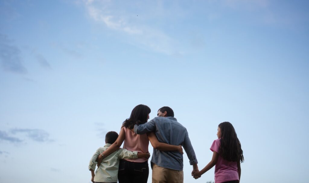 Back view of a latin family of four standing outdoors, embracing and holding hands with the sky in the background.