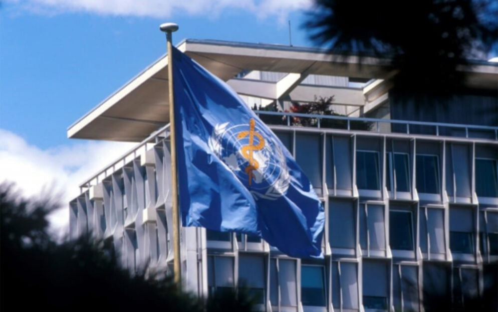 World Health Organization (WHO) flag waving in front of the WHO headquarters building in Geneva, Switzerland on a clear blue sky day.