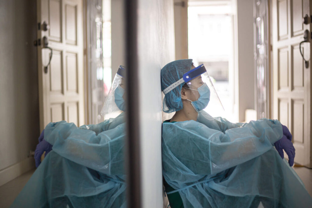 an exhausted medical profession in scrubs, face mask and shield sitting on the floor