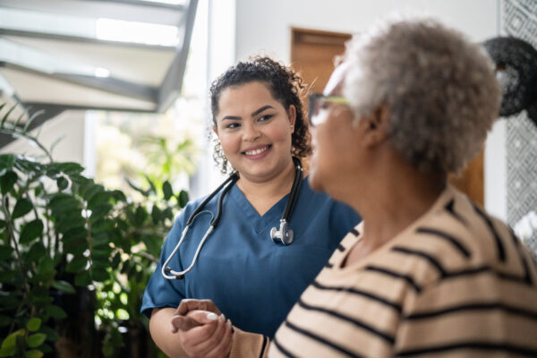 Home caregiver wearing a stethoscope around her neck helping senior woman walking at home