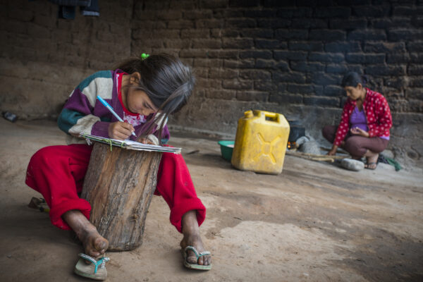 World Chagas Day 2021: All we need is your attention - a young child in a their wolrd country writing on a notepad on top of a log of wood, lady in the background sat at a fire