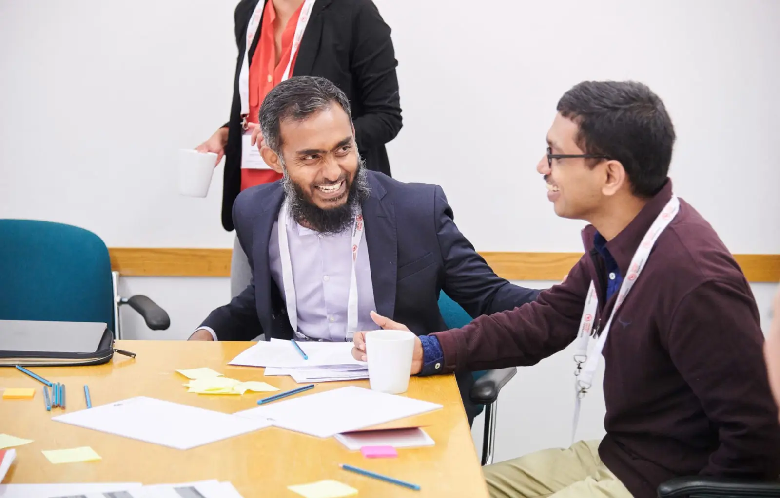 Two men seated at a conference table smiling and talking with each other. One is holding a white coffee mug, and the table is scattered with papers, sticky notes, and pens. A third person stands in the background holding a mug, partially out of frame.