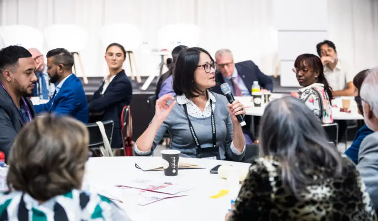 A woman with glasses speaks into a microphone during a roundtable discussion at a conference. She is surrounded by other diverse participants who are listening intently, seated at tables with coffee cups, notebooks, and conference lanyards.