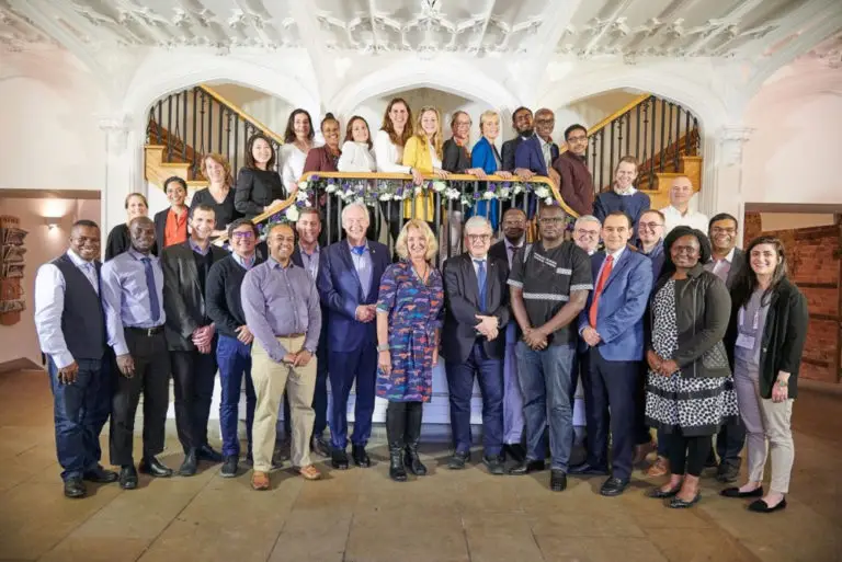 A large, diverse group of professionals poses on a staircase in a grand indoor setting with ornate white ceilings. Some stand on the stairs and others on the floor, all smiling towards the camera. The group are part of the emerging leaders programme.