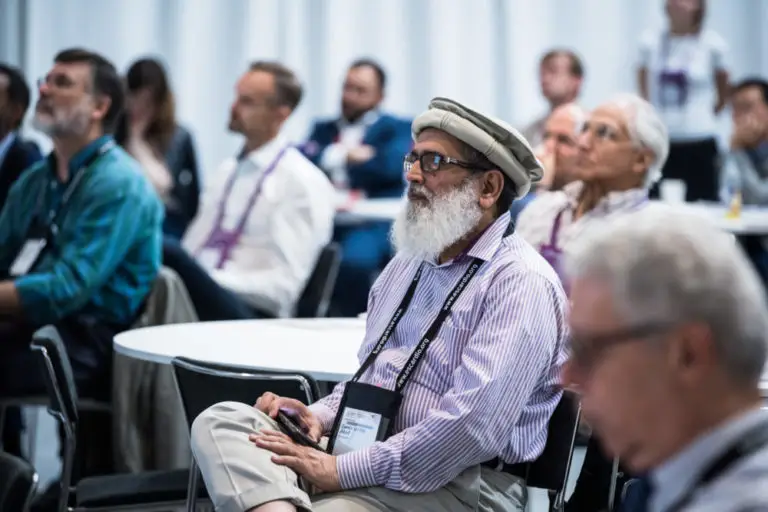 A group of attendees, including an elderly man with a white beard and traditional cap, sit and attentively listen at a conference or seminar. The setting includes round tables and a white backdrop, with people of diverse backgrounds engaged in the session.