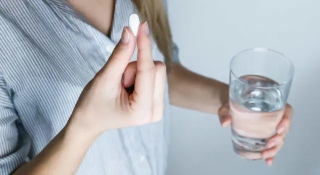 An image of a woman holding a white tablet in her right hand and a glass of water only half filled in her left hand.