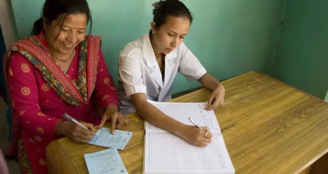 An image of two women in nepal sat registering patients on the sheets of paper infront of them