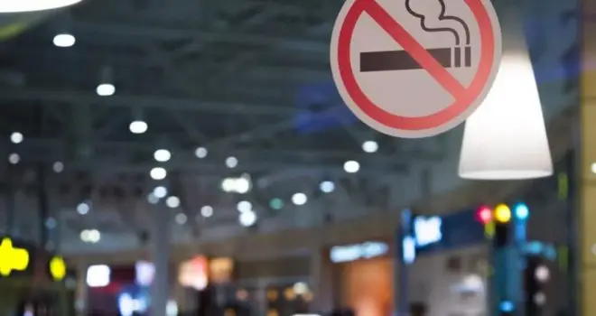 a picture of a no-smoking sign stuck to a window. Pictured behind this are a haze of shop and street lights at night reflecting on the glass window