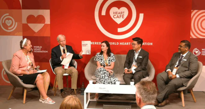 Five panelists seated on stage at a red "Heart Café" backdrop with "Powered by World Heart Federation" branding. From left to right: a woman in a pale pink blazer with a microphone, an older man holding a booklet and gesturing as he speaks, a woman in a patterned dress listening, a young man in a suit, and a man in a business outfit—engaged in a structured discussion around a central white table with water bottles and cups.