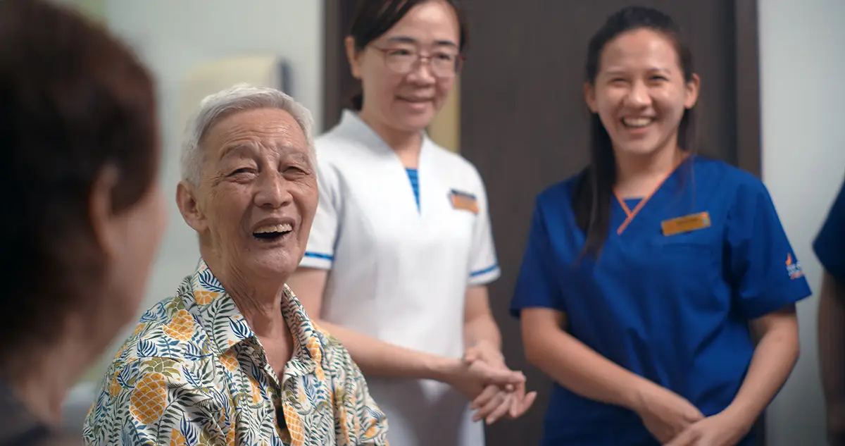 An older man in a pineapple patterned short laughing and smiling at the nurses in the room in white and blue nursing uniform