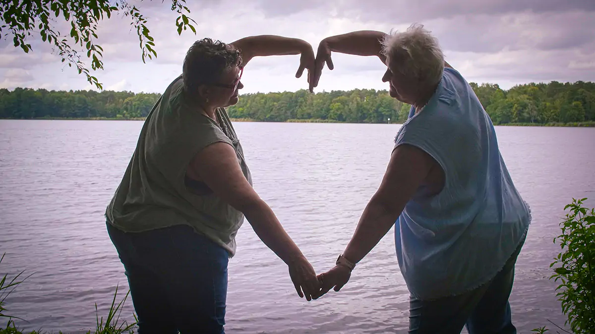 2 women making a heart shape with their arms in front of a lake