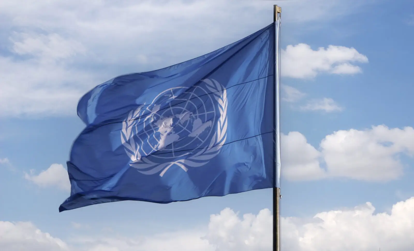 A blue United Nations flag blowing in the wind with a blue sky and clouds in the background