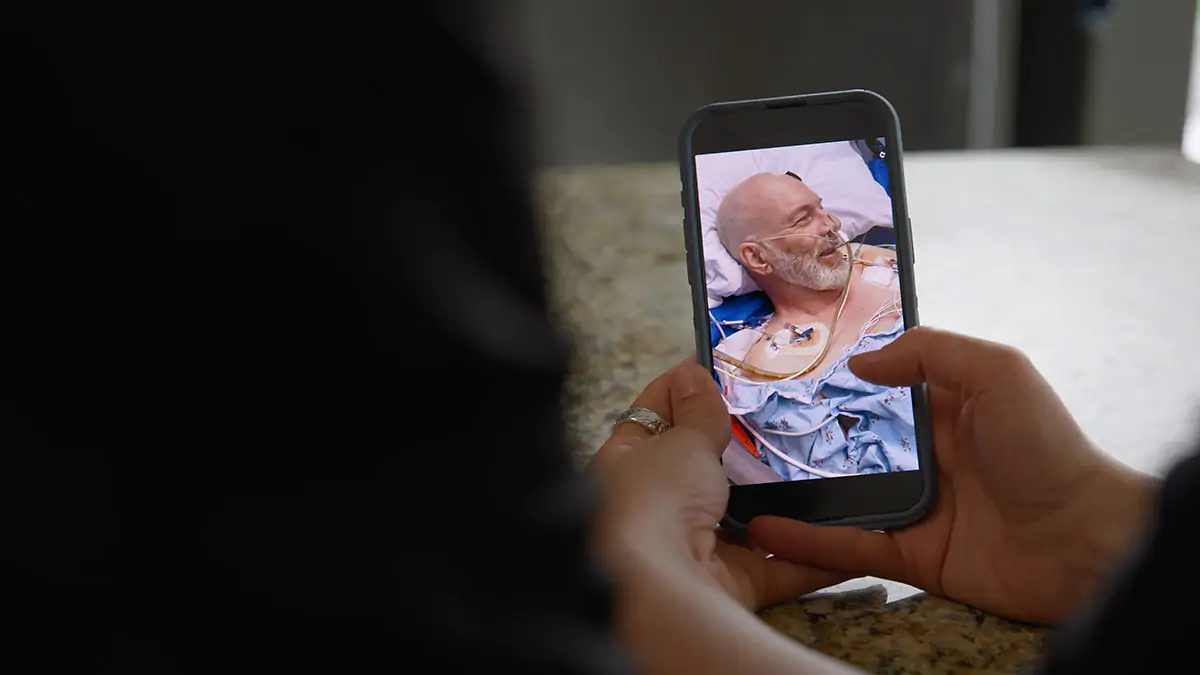 A person holding a phone showing another person with a picture of a man in hospital with tubes coming in and out of him