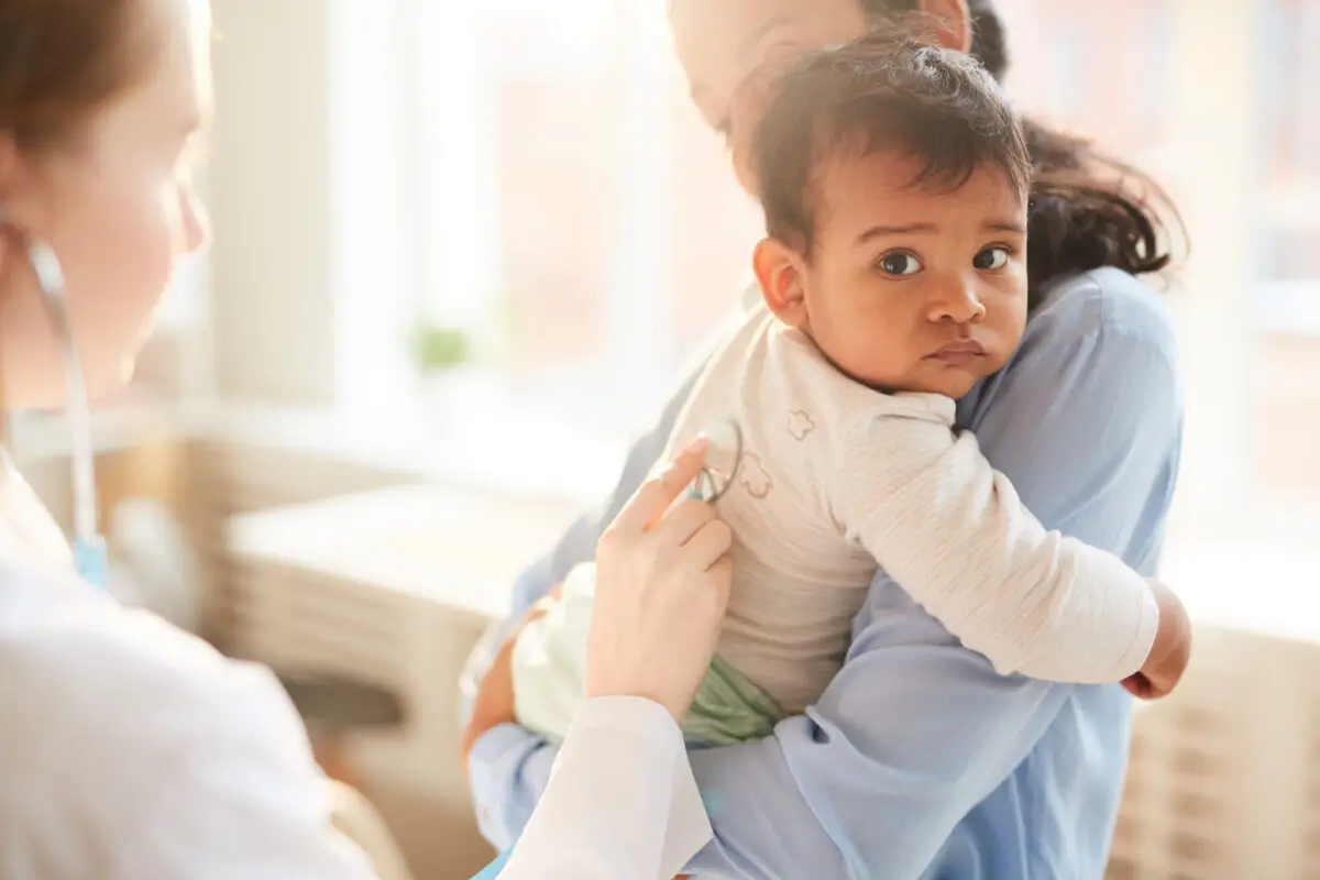 A mother is holding her toddler while a medical professional is using a stethoscope to examine
