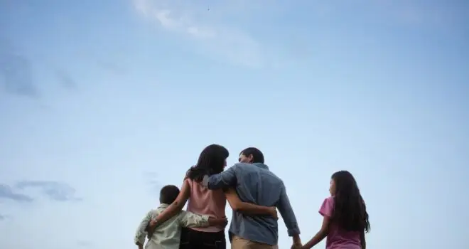 Back view of a latin family of four standing outdoors, embracing and holding hands with the sky in the background.