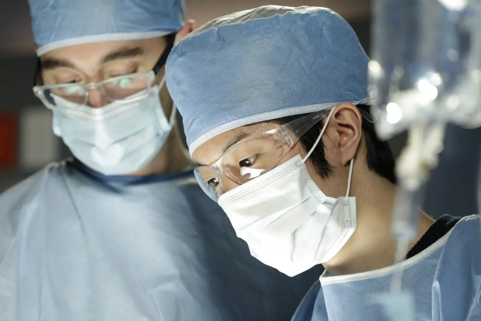 two medical professionals in scrubs, facemak, hats and glasses examining a patient